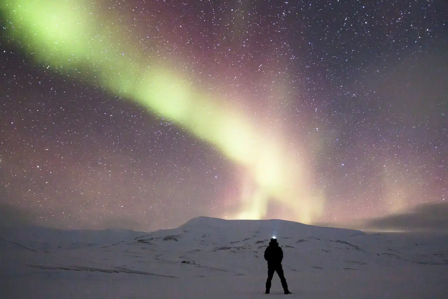 A lone figure with a headlamp under the northern lights on a snow-covered plain.