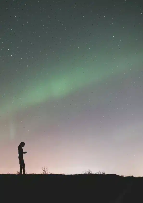 The silhouette of a woman on a ridge as a green aurora crosses the sky.