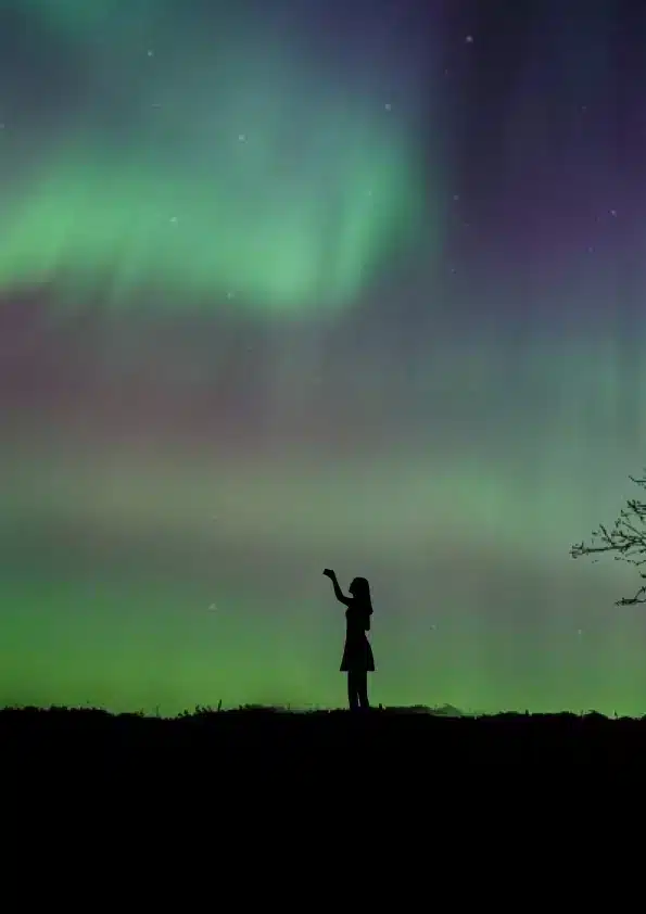 A woman reaching up toward a green and violet aurora.