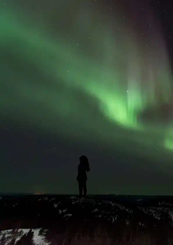 A figure alone on a snowy ridge beneath a wide band of northern lights.