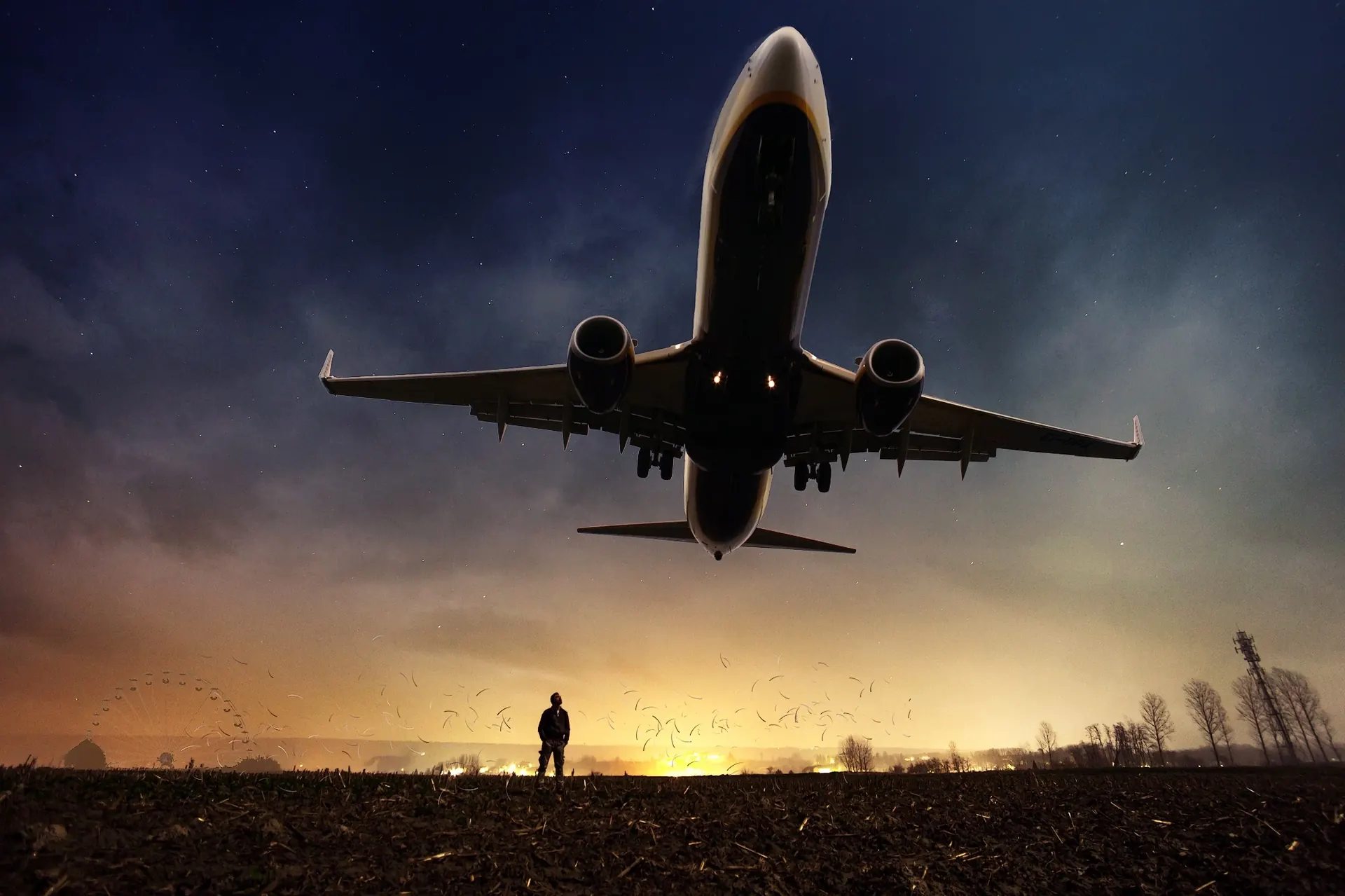 A passenger jet lifting off at dusk while a lone figure watches from the field below